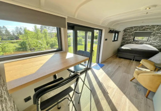 Interior luminoso de Pine Tree Tiny Cottage en Bélgica, con escritorio, cama y vistas al bosque exterior.