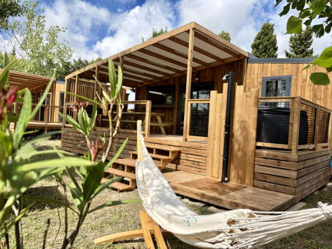 Trælodge ved La Clairiere med overdækket terrasse, hængekøje og grønne planter i forgrunden, solrig dag.