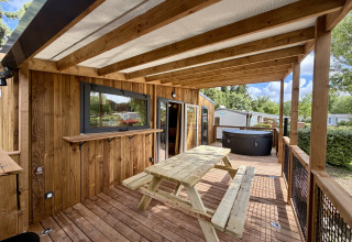 Terrazza coperta in legno con tavolo da picnic e vasca idromassaggio al lodge La Clairiere, Camping Seasonova Ensoya, Francia.
