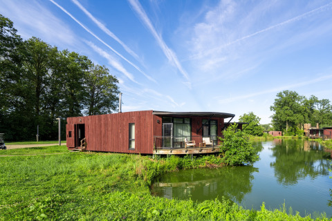 Lodge The Leaf by YourNature aan het meer, omgeven door groen en onder een blauwe lucht met vliegtuigstrepen.
