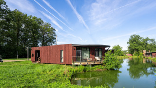 Lodge The Leaf by YourNature aan het meer, omgeven door groen en onder een blauwe lucht met vliegtuigstrepen.