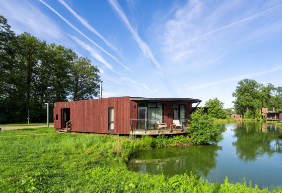Lodge The Leaf by YourNature aan het meer, omgeven door groen en onder een blauwe lucht met vliegtuigstrepen.