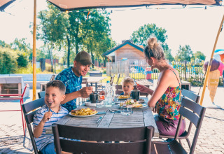 En familie nyder frokost sammen udendørs ved et lodge, børn og forældre smiler og hygger sig.