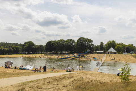 People enjoy a sunny day by a lake with a waterslide and sandy beach at a glamping accommodation.