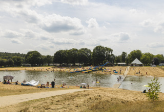 People enjoy a sunny day by a lake with a waterslide and sandy beach at a glamping accommodation.