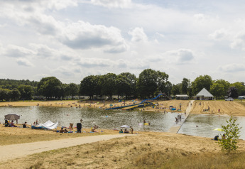 Folk nyder en sommerdag ved en sø med vandrutsjebane og sandstrand på en glampingplads.