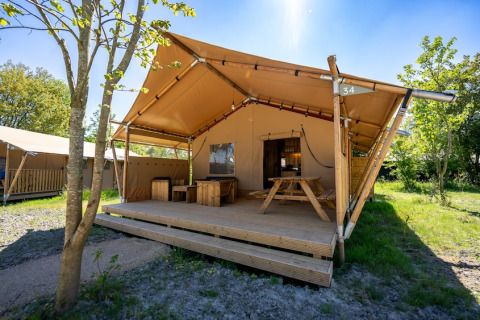 Tenda safari con veranda in legno, tavolo da picnic e panche, immersa nel verde naturale e luminoso.
