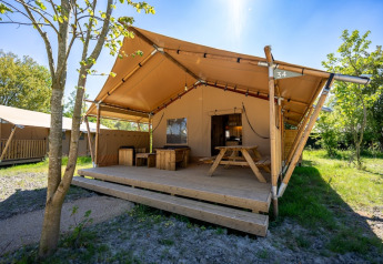Tenda safari con veranda in legno, tavolo da picnic e panche, immersa nel verde naturale e luminoso.