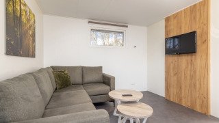 Sala de estar en Chalet Comfort 4 con sofá gris en L, dos mesas redondas y televisor en la pared.