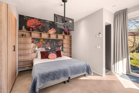 Modern bedroom with a floral mural, double bed, wood shelving, and ceiling-mounted TV in natural light.