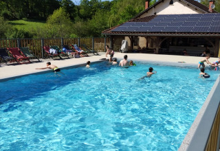 Buitenzwembad met mensen, ligstoelen en huis met zonnepanelen bij Domaine Papillon, Occitanie, Frankrijk.