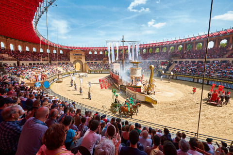 Spettatori guardano uno spettacolo romano con carri e nave in una grande arena all'aperto.