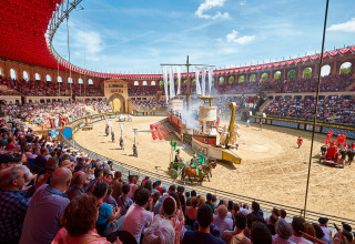Spettatori guardano uno spettacolo romano con carri e nave in una grande arena all'aperto.