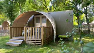 Tenda safari Coco Sweet con veranda in legno, circondata da erba verde e alberi in un campeggio tranquillo.