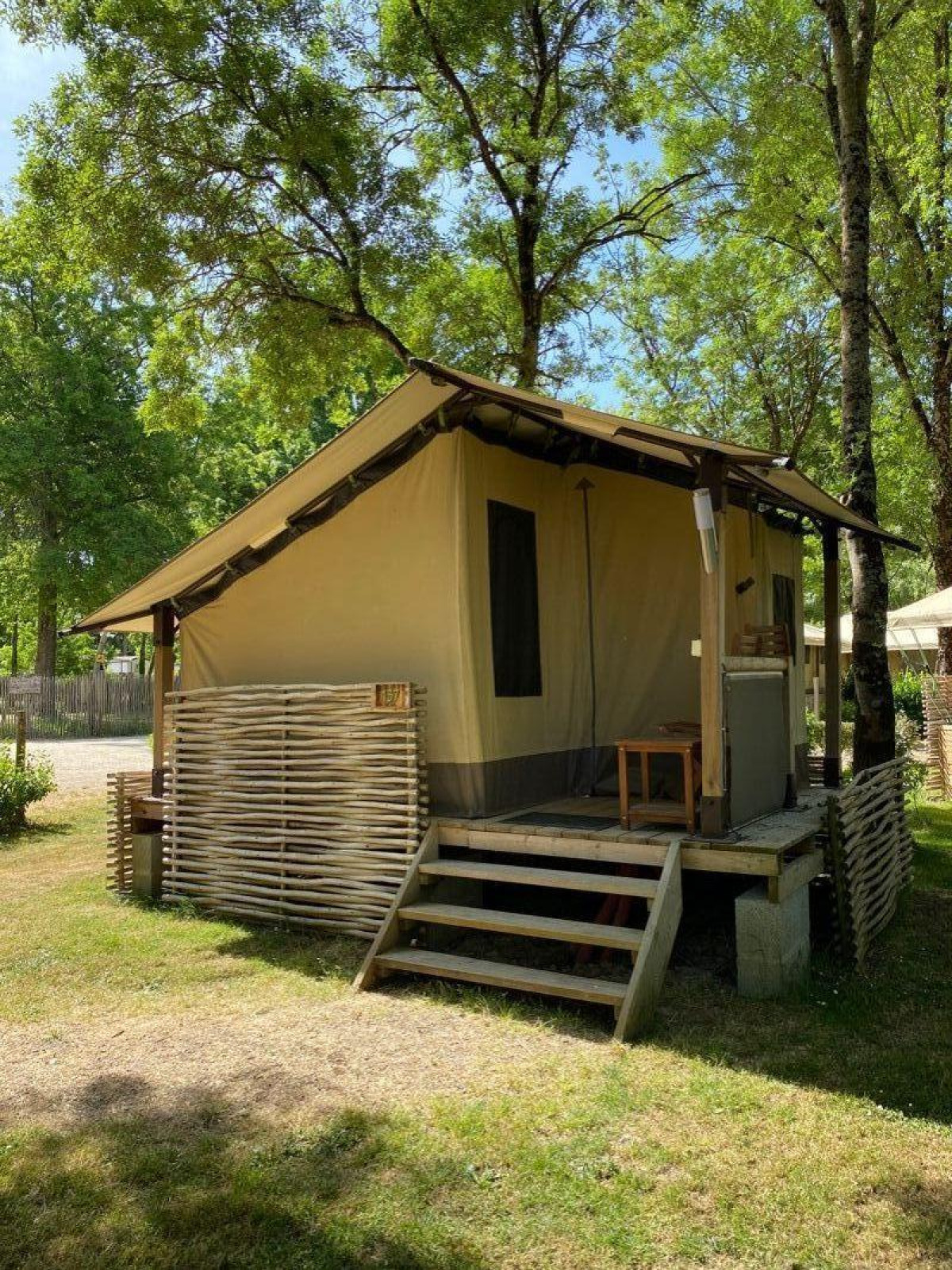 Safaritent Lodge Balearic met houten veranda, omgeven door groene bomen en natuur op een zonnige dag.