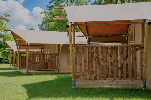 Safari tents with wooden decks on grass at Baalse Hei, Belgium, surrounded by trees and blue sky.