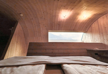 Innenraum des Ufogel-Naturhauses in Österreich mit gewölbter Holzdecke, Bett und Bergblick.