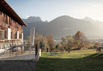 Blick auf das Ufogel-Naturhaus in Österreich mit Bergen, rustikalen Gebäuden und sonniger Wiese.