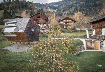 Moderne Naturhaus Ufogel in Österreich, von traditionellen Holzhäusern und Waldlandschaft umgeben.