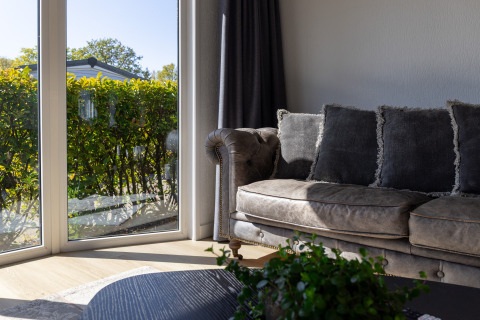 Living room at Wilbrinkchalet, De Boshoek, Netherlands, with sofa, large windows and green hedge outside.