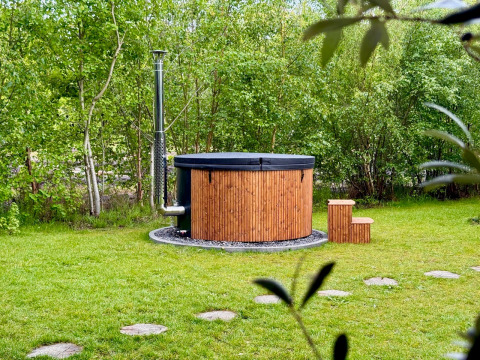 Vasca idromassaggio esterna in legno con camino, immersa nel verde e circondata da alberi presso un lodge.