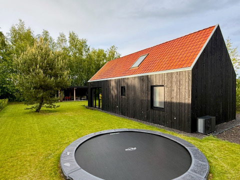 Moderne zwarte boshut met rood dak, grasveld, ingegraven trampoline en omringd door groene bomen.