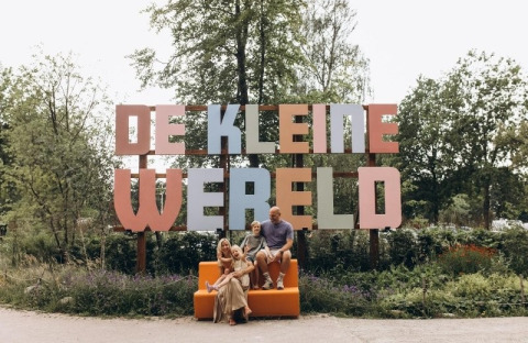 Familia sentada ante el letrero 'De Kleine Wereld' en un camping rodeado de naturaleza en Groningen, Países Bajos.