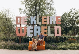 Familie sitzt vor dem großen Schild 'De Kleine Wereld' auf einem Ferienpark in Groningen, Niederlande im Grünen.