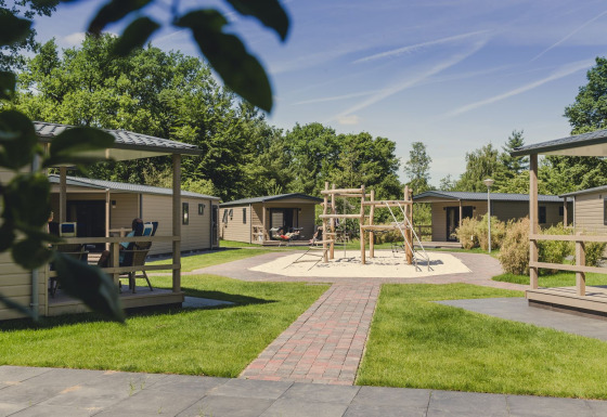Lodge moderno Bungalette Comfort con porche, zona de juegos central y áreas verdes en un día soleado.
