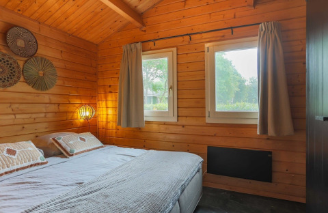 Cozy lodge bedroom with wooden walls, a double bed, two windows, soft lighting, and nature outside.