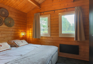 Cozy lodge bedroom with wooden walls, a double bed, two windows, soft lighting, and nature outside.