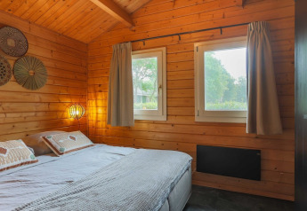 Cozy lodge bedroom with wooden walls, a double bed, two windows, soft lighting, and nature outside.