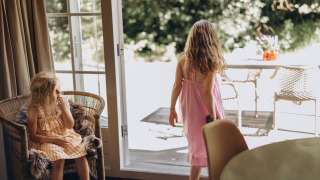 Two girls in summer dresses, one sitting and one walking outside, at a lodge with an open patio door.
