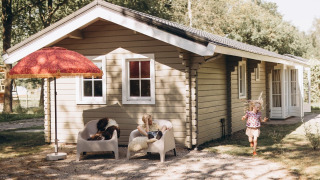 Due bambini giocano davanti a un rifugio accogliente con pareti beige, sedie e un grande ombrellone rosso.