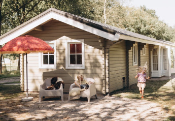 Two children play outside a cozy lodge with beige wooden walls, armchairs, and a large red umbrella.