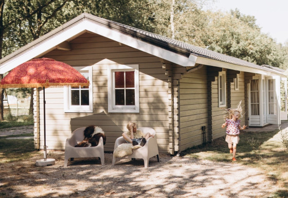 Twee kinderen spelen voor een gezellige lodge met stoelen en een grote rode parasol in de tuin.