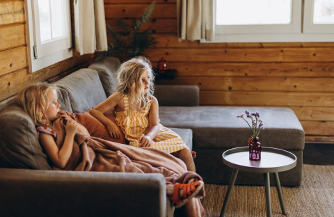 Twee kinderen zitten op een grijze zetel onder een deken in een houten lodge, met tafeltje en bloemenvaas.