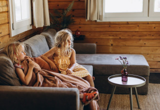 Twee kinderen zitten op een grijze zetel onder een deken in een houten lodge, met tafeltje en bloemenvaas.