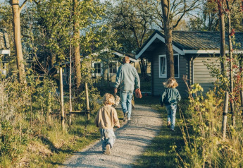 Family walking on a path to Swedish Village House at Camping De Kleine Wereld in the Netherlands, surrounded by trees.