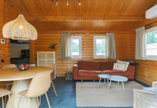 Interior acogedor de una cabaña con paredes de madera, mesa redonda, sofá, alfombra y ventanas con cortinas.