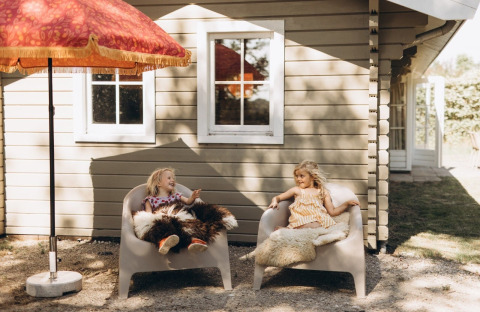 Two children sit and chat on chairs covered with fur outside a lodge under a red umbrella in sunlight.