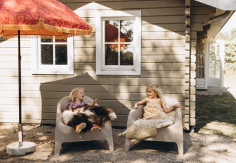 Two children sit and chat on chairs covered with fur outside a lodge under a red umbrella in sunlight.