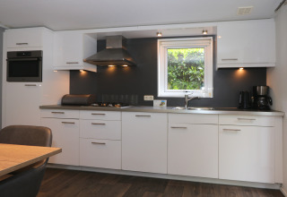 Moderne Küche mit weißen Schränken, Fenster, Geräten und Esstisch in L-Pavilion 6, De Achterhoek, Niederlande.
