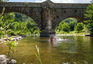 Menschen baden im Fluss unter einer Steinbrücke inmitten grüner Natur im Flower Camping Le Pont du Tarn.