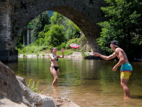 Twee jongeren spelen frisbee in een rivier onder een stenen brug bij Flower Camping Le Pont du Tarn in Occitanië, Frankrijk.