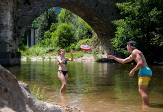 To unge leger med en frisbee i en flod under en stenbro ved Flower Camping Le Pont du Tarn i Occitanie, Frankrig.