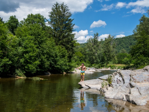 Man stands on rocks by a river surrounded by lush trees at Flower Camping Le Pont du Tarn in Occitanie, France.