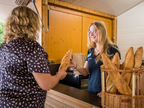 Vrouw koopt vers stokbrood bij een glimlachende verkoopster op Flower Camping Le Pont du Tarn in Frankrijk.