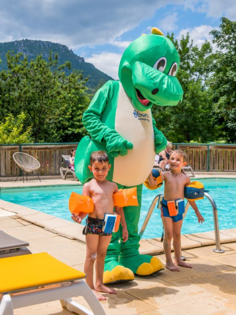 Two children with armbands by the pool and a large dinosaur mascot at Flower Camping Le Pont du Tarn in France.