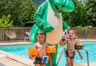 Twee kinderen met zwembandjes bij het zwembad en een grote dinosaurusmascotte bij Flower Camping Le Pont du Tarn.